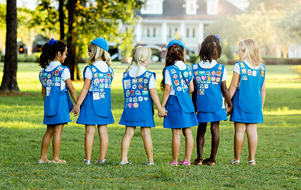 group of daisy girl scouts holding hands in uniform blue apron vest with patches and badges
