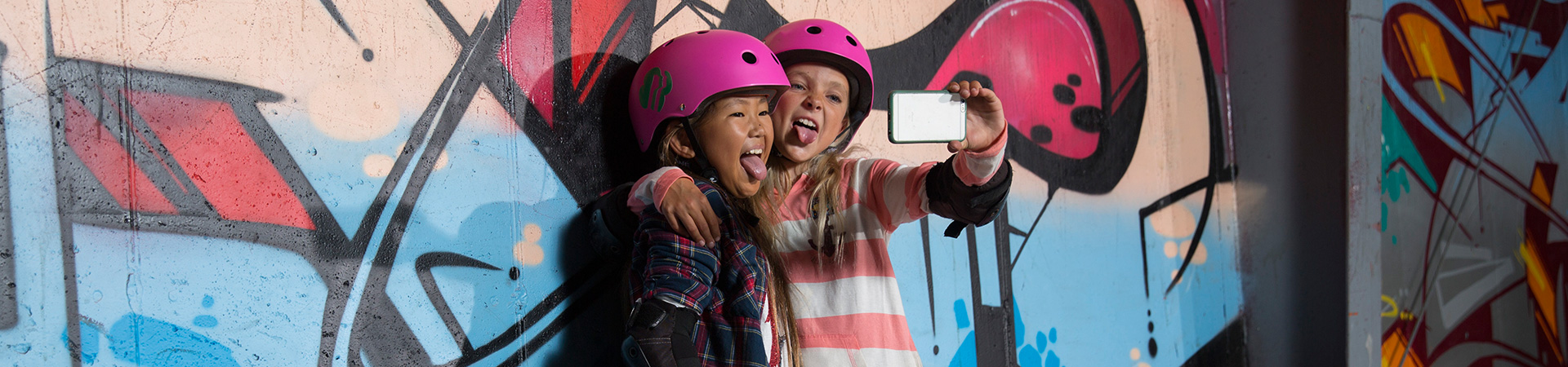  two girls wearing pink skateboarding helmets taking selfie against graffiti wall 